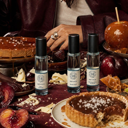 A woman in a burgundy leather jacket sits at a table surrounded by caramel apples, tart, plums, and three small fragrance bottles from Scented Objects. She holds a caramel apple near the camera, with the products arranged in front of her.